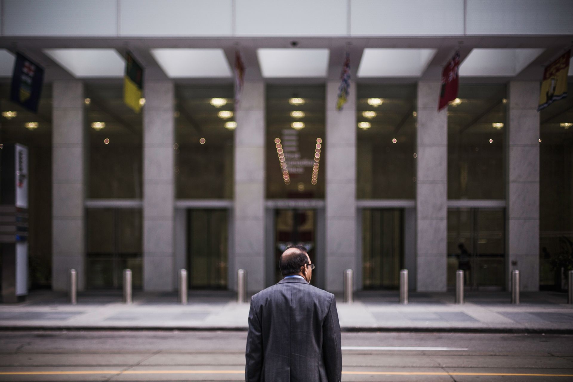 Man in suit stands on street facing large modern building with tall glass windows and flags.