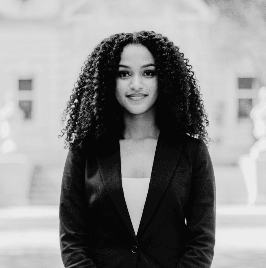 Black and white portrait of a person with curly hair in a blazer, standing outdoors.