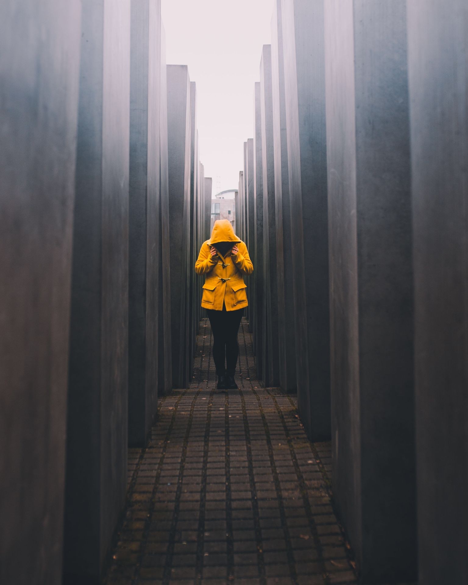 Person in a yellow coat walking between tall, narrow concrete columns on a patterned floor.